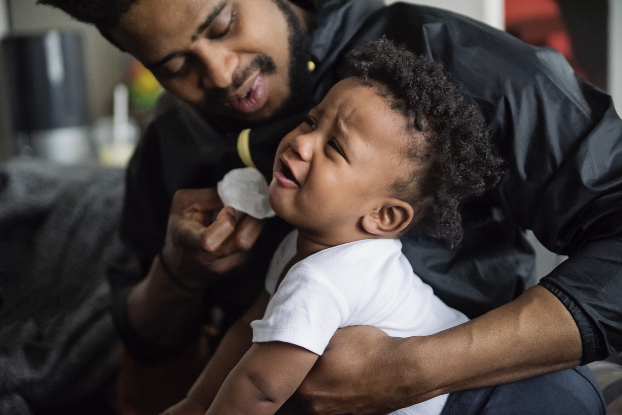 Father wiping the face of unhappy baby boy. Dad is in thirties, baby is one year’s old. Horizontal waist up indoors shot in natural light. Soft focus. Some copy space.